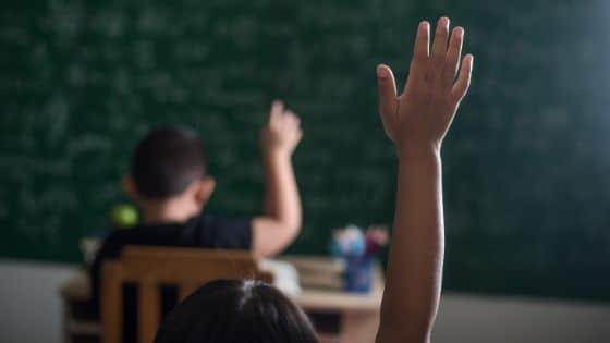 Menina levanta braço em pedido para falar na sala de aula. Ao fundo, outra criança, próxima a um quadro negro, também levanta o braço.