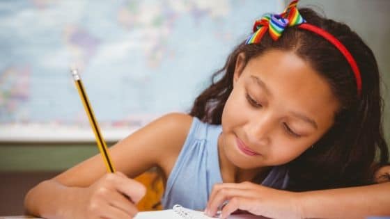 Menina escrevendo de lápis no caderno. Ao fundo, há um mapa-múndi.